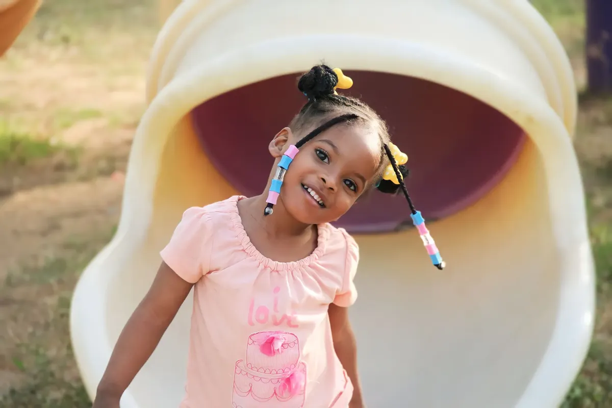 A joyful child with braids playing outdoors in a pink shirt and tube slide.