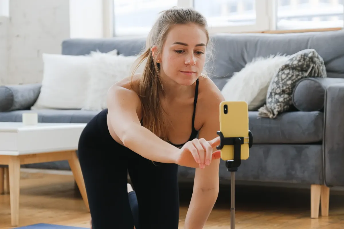 若き女性がアクティウェアを着て、自宅でスマートフォンを三脚に固定してヨガの練習をログしている。